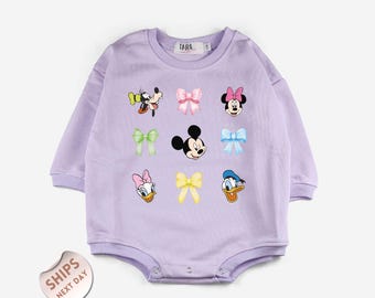 May include: A light purple baby onesie with long sleeves. The onesie features cartoon character graphics including Mickey Mouse, Minnie Mouse, Donald Duck, and Daisy Duck, along with various bows. The brand name "TARA" is printed on the top.