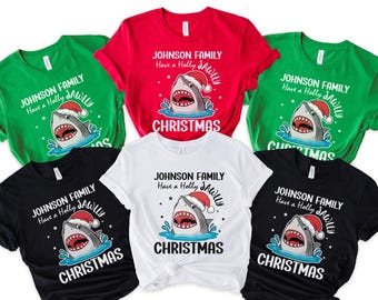 May include: A collection of Christmas-themed t-shirts in various colors, including green, red, black, and white. Each shirt features a shark wearing a Santa hat, with the text "JOHNSON FAMILY Have a Holly JAWLLY CHRISTMAS".