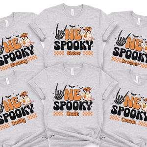 A collection of gray t-shirts with Halloween-themed designs, including a skeleton hand, a ghost, and the text "NE SPOKEY" and "Sister" printed on them.