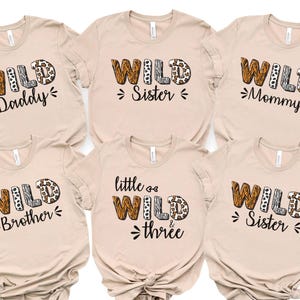 May include: Six beige t-shirts with the word "WILD" in a mix of animal print patterns. Each shirt has a different family member title: Daddy, Sister, Mommy, Brother, Little & Three, and Sister. The shirts are casual wear.