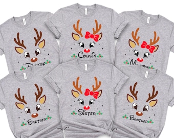 May include: A collection of six light gray short-sleeve t-shirts, each featuring a graphic of a reindeer face with brown antlers, a red nose, and black eyes. Some reindeer have a red bow. Text includes Daddy, Cousin, Mommy, Brother, and Sister.