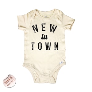 May include: Cream-colored baby onesie with the text "NEW in TOWN" in distressed black lettering. The onesie has short sleeves and a snap closure at the bottom. The Gerber brand logo is visible.