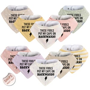 May include: A collection of baby bibs in various pastel colors, each featuring the phrase "THESE FOOLS PUT MY CAPE ON BACKWARDS" in bold black text. The bibs have a triangular shape with snap closures and are made of soft fabric.