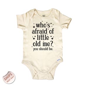 May include: Cream-colored baby bodysuit with the text "who's afraid of little old me? you should be." in black. The bodysuit is short-sleeved and has a snap closure.