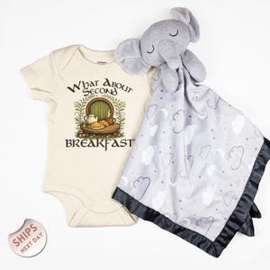May include: A cream-colored baby onesie with the text "What About Second Breakfast" and a graphic of a hobbit door and food. A gray elephant plush toy rests on a gray and white cloud-patterned security blanket with a black satin trim.