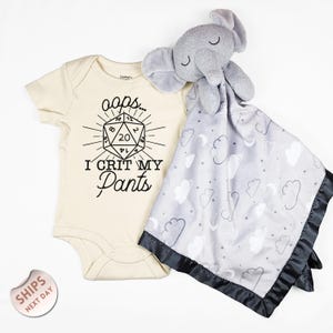 May include: A cream-colored baby onesie with the text "oops... I crit my pants" and a D20 graphic. A gray plush elephant toy and a gray and white cloud-patterned security blanket with a black satin trim are also included.