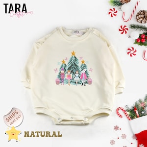 May include: Cream-colored baby romper with a crew neck and long sleeves. The front features a colorful graphic of a Christmas tree scene with characters. The text "TARA Outfit" is visible. The word "NATURAL" is also visible.