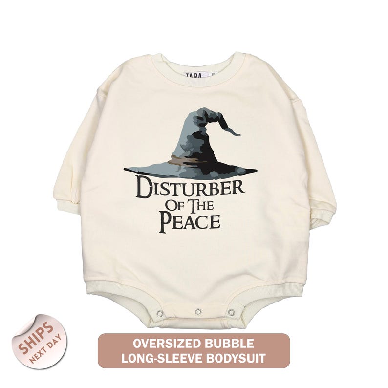 An oversized white long-sleeved bodysuit with a graphic of a wizard's hat and the text "Disturb of the Peace" printed on it.