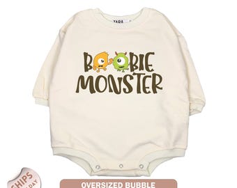 May include: Cream-colored, long-sleeved baby bodysuit with the text "BOOBIE MONSTER" in brown. The design includes cartoon monster graphics in orange and green. The bodysuit has snap closures and a round neckline.