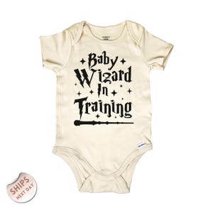 May include: Cream-colored baby onesie with the text "Baby Wizard In Training" in black, along with stars, lightning bolts, and a wand design. The onesie is made of soft material and has short sleeves.