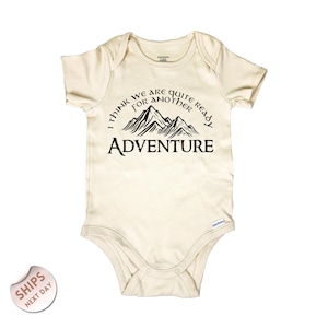 May include: Cream-colored baby onesie with short sleeves. The front features a black graphic of mountain peaks and the text "I THINK WE ARE QUITE READY FOR ANOTHER ADVENTURE."