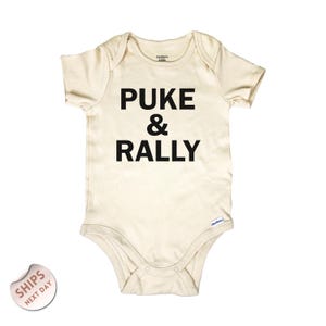 May include: Cream-colored baby onesie with the words "PUKE & RALLY" printed in bold black letters. The onesie is short-sleeved and has snap closures at the bottom. The Gerber brand logo is visible.