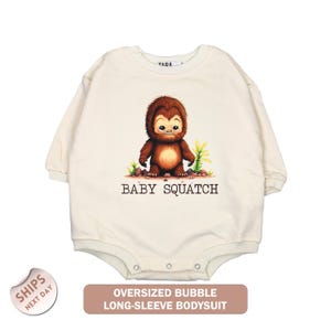 May include: Cream-colored, long-sleeved baby bodysuit with a cartoon illustration of a brown Bigfoot character. The text "BABY SQUATCH" is below the illustration. The bottom of the bodysuit has snap closures. The text "OVERSIZED BUBBLE LONG-SLEEVE BODYSUIT" is at the bottom.