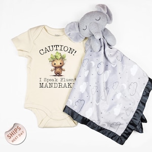 May include: A cream-colored baby onesie with the text "CAUTION! I Speak Fluent MANDRAK!". A gray plush elephant and a gray and white cloud-patterned security blanket with a black satin trim are also in the image.