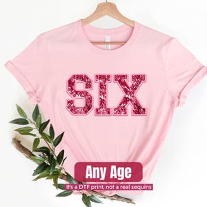 May include: A light pink t-shirt with the word "SIX" in a sequin-like design. The shirt has rolled-up sleeves and is hanging on a wooden hanger. A banner at the bottom reads "Any Age" with the text "It's a DTF print, not a real sequins".