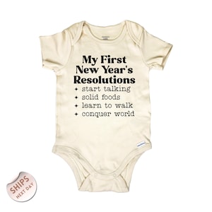 May include: Cream-colored baby onesie with the text "My First New Year's Resolutions" and a list of goals in black. The goals include "start talking," "solid foods," "learn to walk," and "conquer world."