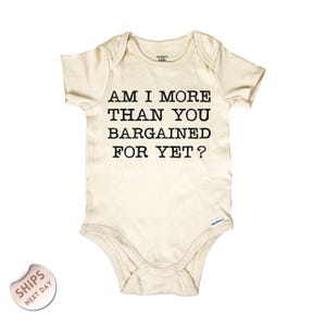 May include: Cream-colored baby bodysuit with the text "AM I MORE THAN YOU BARGAINED FOR YET?" printed in black. The bodysuit has short sleeves and snap closures at the bottom.