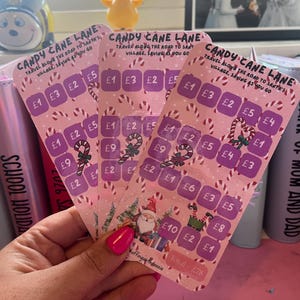 Può includere: Tre biglietti gratta e vinci rosa Candy Cane Lane a tema natalizio. Ogni biglietto presenta un design a bastoncino di zucchero e il testo "Candy Cane Lane, Viaggia lungo la strada per il villaggio di Babbo Natale, Risparmiando mentre vai."