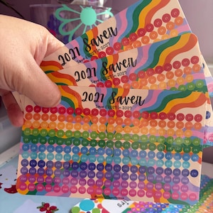 Könnte beinhalten: Ein Set rechteckiger 2021 Saver-Karten mit regenbogenfarbenem Wellenhintergrund. Jede Karte zeigt den Text 2021 Saver und Save £2027 in 2027 sowie Reihen kreisförmiger Symbole, die mit verschiedenen Pfund-Beträgen zur Sparverfolgung beschriftet sind.