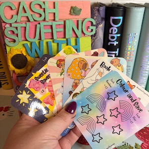 May include: A hand displaying a selection of illustrated cards for a cash stuffing system. The cards feature rainbows, stars, and cartoon characters. A wooden sign in the background reads "CASH STUFFING WITH".