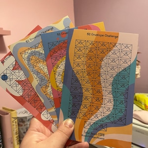Può includere: Quattro tracker colorati per la sfida delle 52 buste con un totale risparmiato di £ 1378. I tracker sono stampati con uno sfondo ondulato e hanno un contorno nero di una busta con un cuore all'interno. I tracker sono stampati su carta bianca.
