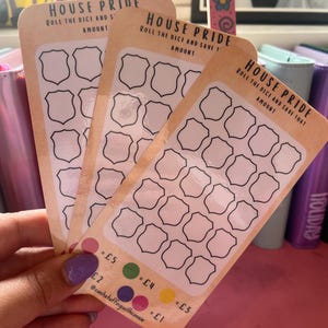 Może przedstawiać: Trzy beżowe karty z napisem "HOUSE PRIDE" i "ROLL THE DICE AND SAVE THAT AMOUNT". Każda karta ma siatkę pustych kształtów tarcz i kolorowych kół z kwotami od £1 do £5. Karty są trzymane w dłoni.