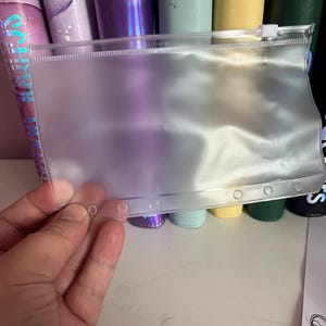Puede incluir: Una bolsa transparente de plástico con cremallera y seis agujeros en el borde inferior. La bolsa está encima de seis carpetas de diferentes colores. Las carpetas son de color morado, verde, amarillo y negro.