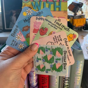 May include: A hand holding several colorful cards with various designs and text, including "Daisy of", "Mududdles", "Peppa", "Food crush", and "DinoSaur Rawr". The cards are part of a savings challenge, with text like "Save 20p for each section you colour in".