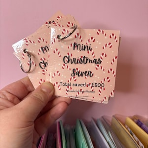 Può includere: Un mini salvadanaio di Natale rosa e bianco con un motivo a bastoncino di zucchero e il testo "Mini Christmas Saver" e "Total saved = £600" con l'hashtag #cashstuffingwithcassie.