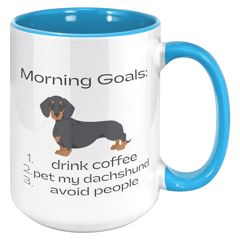 May include: White coffee mug with a blue rim and handle. The mug has a black and tan dachshund illustration and the text "Morning Goals: 1. drink coffee 2. pet my dachshund 3. avoid people".