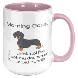 May include: White coffee mug with a pink rim and handle. The mug has a black and tan dachshund illustration and the text "Morning Goals: 1. drink coffee 2. pet my dachshund 3. avoid people".