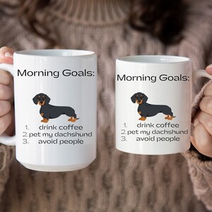 May include: Two white coffee mugs with black handles. Both mugs feature a black and tan dachshund illustration and the text "Morning Goals:" followed by a list of three goals: "drink coffee", "pet my dachshund", and "avoid people".