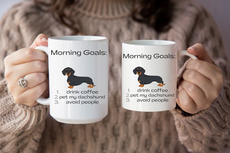 May include: Two white coffee mugs with black handles. Both mugs feature a black and tan dachshund illustration and the text "Morning Goals:" followed by a list of three goals: "drink coffee", "pet my dachshund", and "avoid people".