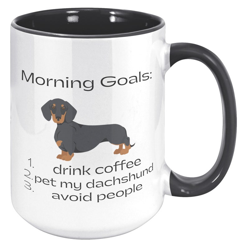 May include: White coffee mug with a black rim and handle. The mug has a black and tan dachshund illustration and the text "Morning Goals: 1. drink coffee 2. pet my dachshund 3. avoid people".