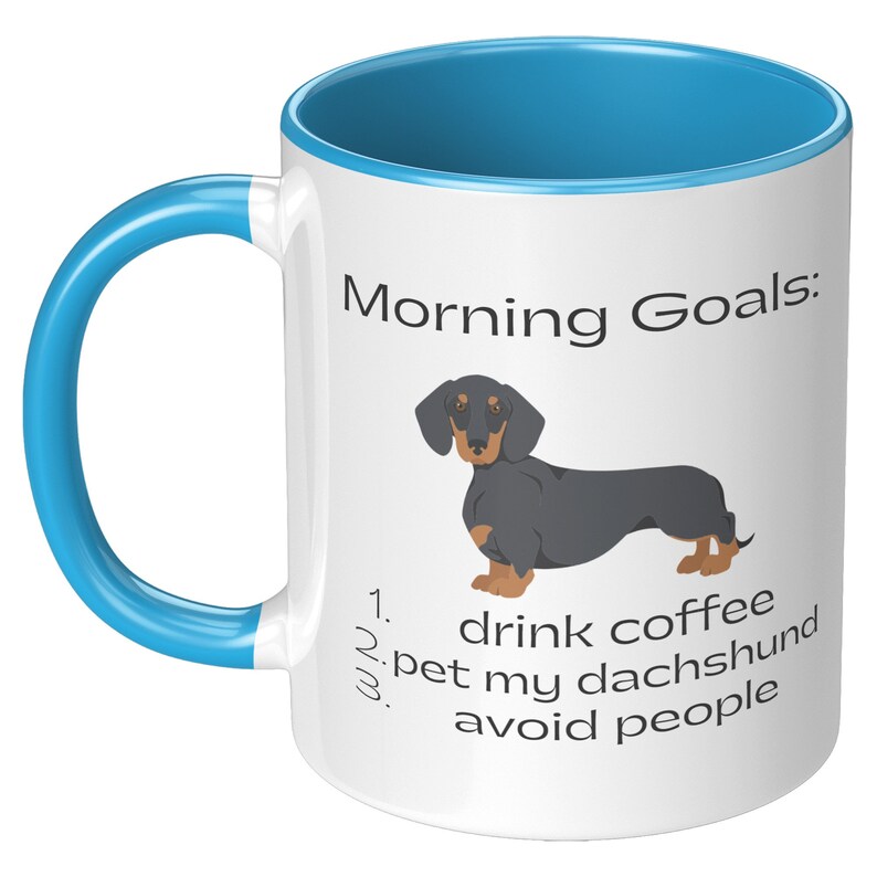 May include: White ceramic coffee mug with a blue rim and handle. The mug has a black and brown dachshund illustration and text that reads "Morning Goals: 1. drink coffee 2. pet my dachshund 3. avoid people".