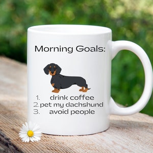 May include: White coffee mug with a black and tan dachshund illustration. The mug reads "Morning Goals: 1. drink coffee 2. pet my dachshund 3. avoid people"