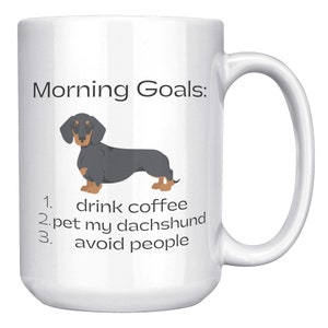 May include: White coffee mug with a black and tan dachshund illustration and the text "Morning Goals:" 1. drink coffee 2. pet my dachshund 3. avoid people