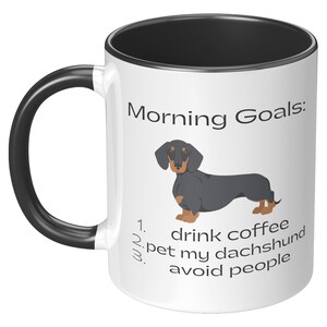 May include: White coffee mug with a black rim and handle. The mug has a black and tan dachshund illustration and the text "Morning Goals: 1. drink coffee 2. pet my dachshund 3. avoid people".
