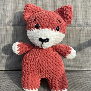 May include: A crocheted plush fox toy with a red body, white face, and black nose. The toy has large black eyes and is sitting on a gray textured surface.