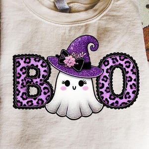 May include: A beige crewneck sweatshirt featuring a graphic of a white ghost wearing a purple glittery witch hat with a black bow and pink flowers. The letters B and O are printed in a purple leopard print pattern with black scalloped borders.