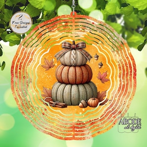 Op de afbeelding: Een decoratieve windspinner met herfstthema, met een stapel van drie pompoenen met een geruite strik, omringd door bladeren en eikels. De tekst "2 Free Designs Included" en "ABODE art digital" zijn inbegrepen. De kleuren zijn oranje, bruin en groen.