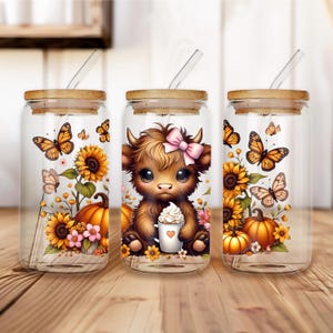 May include: Three glass tumblers with bamboo lids and glass straws. Each tumbler features a different design: butterflies, sunflowers, pumpkins, and a Highland cow with a pink bow holding a coffee cup. The tumblers are on a wooden surface.