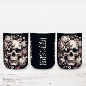 May include: White ceramic mug featuring a detailed skull design surrounded by pink and cream-colored flowers. The mug has a black background and the name "ISABELLA" is printed vertically on the back.