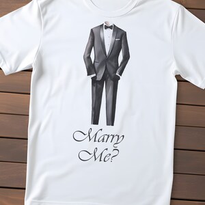 以下が含まれることがあります： 黒いタキシードを着た男性のグラフィックと「Marry Me?」の文字が前面にプリントされた白いTシャツです。
