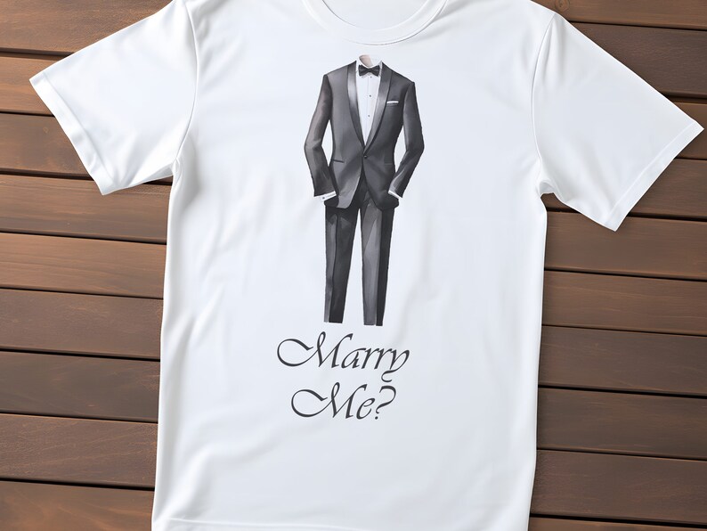 以下が含まれることがあります： 黒いタキシードを着た男性のグラフィックと「Marry Me?」の文字が前面にプリントされた白いTシャツです。