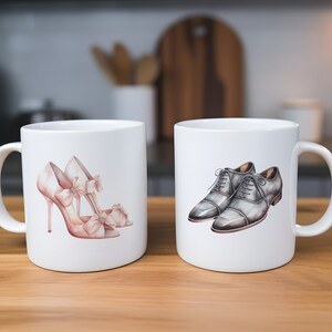 May include: Two white ceramic mugs, each with a shoe design. One has pink high heels with bows, the other features grey dress shoes. The mugs are on a wooden surface, with a blurred kitchen background.