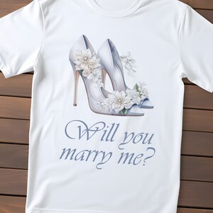 May include: White t-shirt with a watercolour illustration of white high heels decorated with flowers. The text "Will you marry me?" is written in a script font below the heels. The shirt is made of a soft, lightweight material.