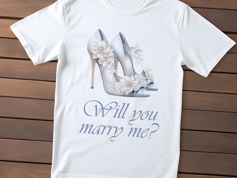 May include: White t-shirt with a watercolour illustration of white high heels decorated with flowers. The text "Will you marry me?" is written in a script font below the heels. The shirt is made of a soft, lightweight material.