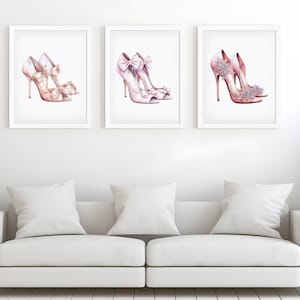 May include: Three framed prints showcasing watercolour illustrations of pink high heels. Each print depicts a different pair of shoes, with details like bows and floral accents. The artwork is displayed above a white sofa with cushions.