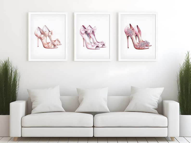 May include: Three framed prints showcasing watercolour illustrations of pink high heels. Each print depicts a different pair of shoes, with details like bows and floral accents. The artwork is displayed above a white sofa with cushions.